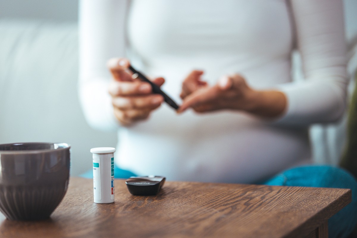 A woman, pregnant and holding a coffee cup with a stirrer in it. She sits at a table with a bottle of supplements and a card.