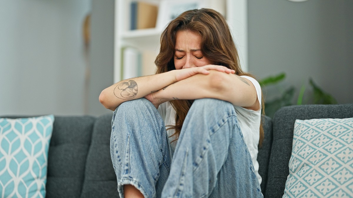 A woman wearing jeans, sitting on a couch with her hand on her face and crying.