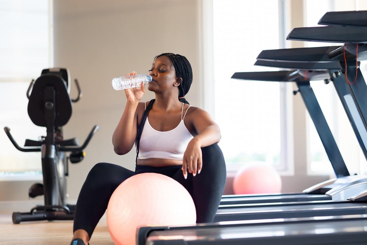 Woman drinking water in gym setting with exercise equipment.