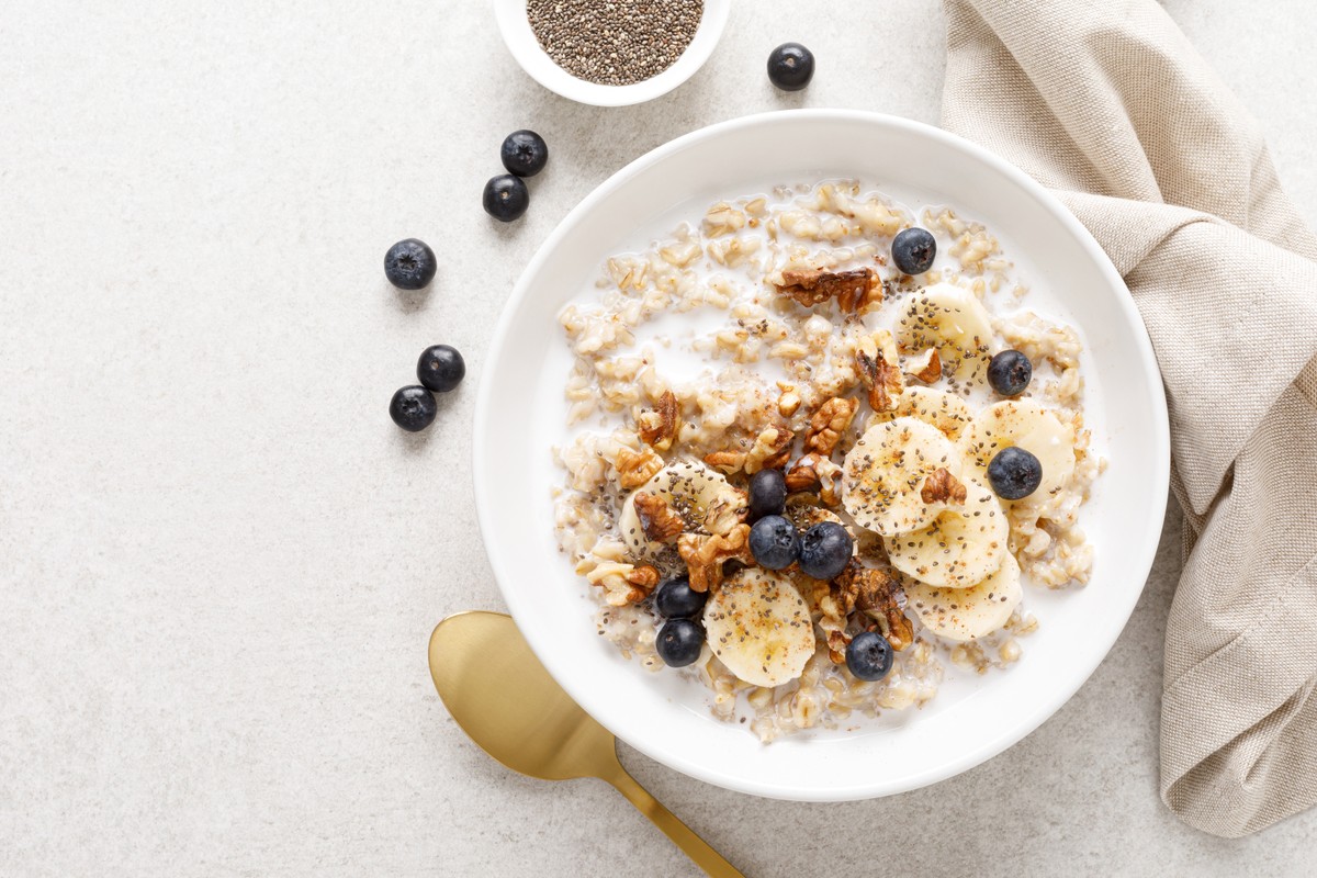 Bowl of oatmeal with banana slices and a spoon, accompanied by blueberries on a white surface.