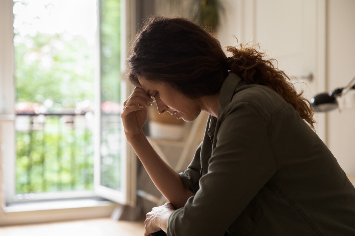Woman in a jacket with her hand on her head, sitting at a table and looking out the window.
