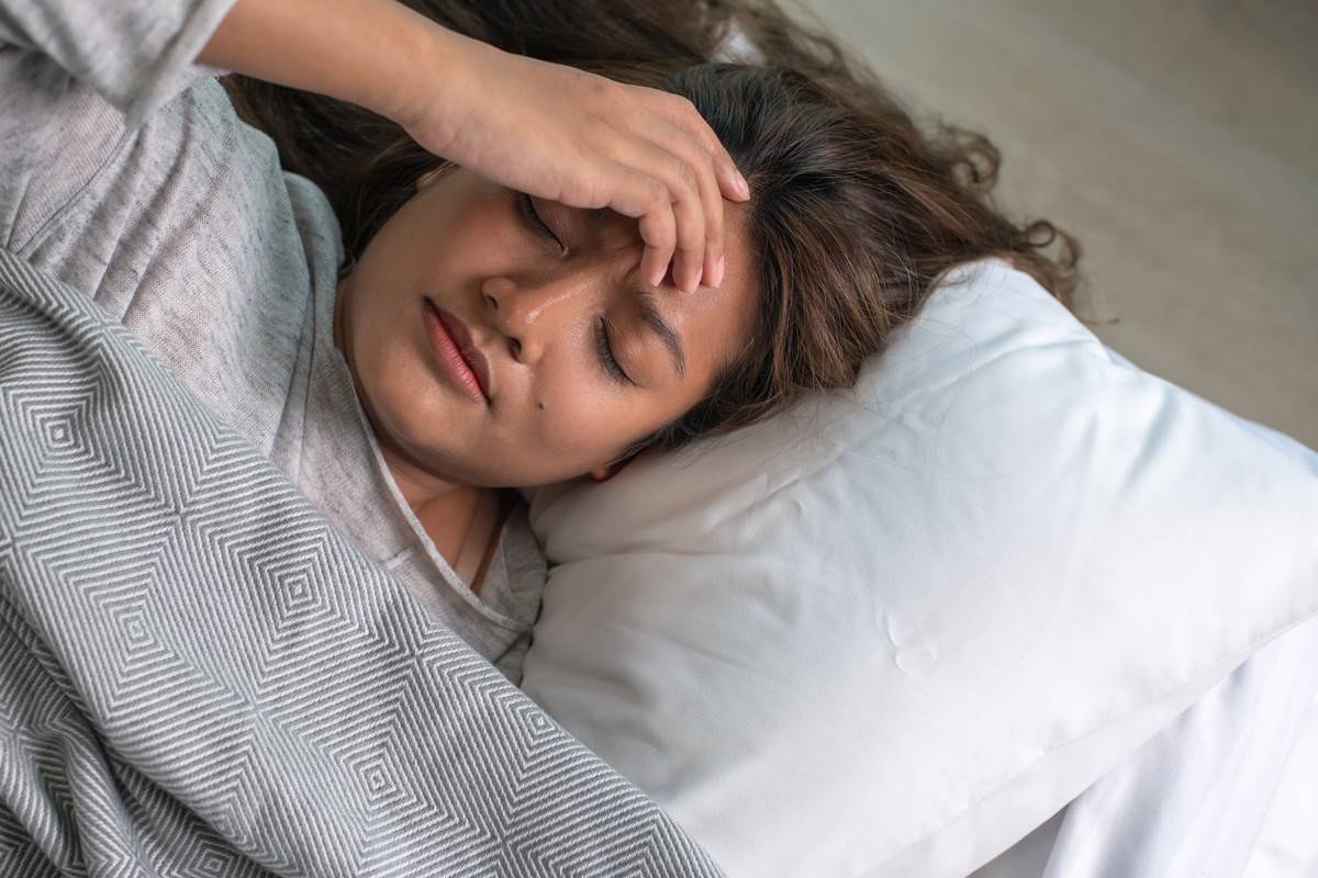 Woman lying in bed, hand on her forehead with a concerned or possibly nauseous expression.
