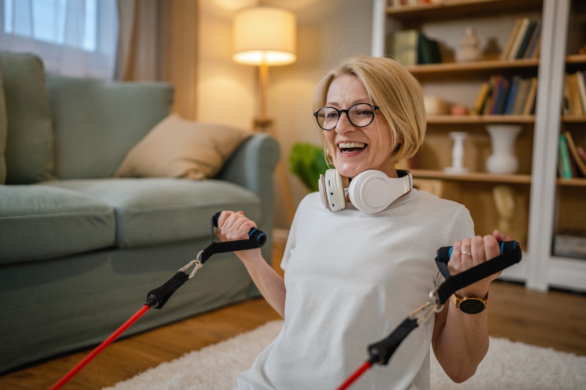 Woman in white shirt holding two weights and smiling, with a living room setting in the background.