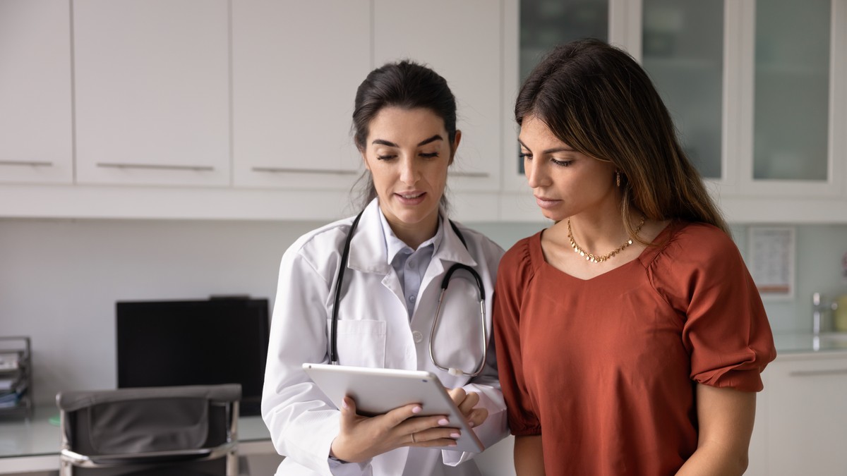 Doctor in white coat examining tablet, standing next to patient who looks concerned.