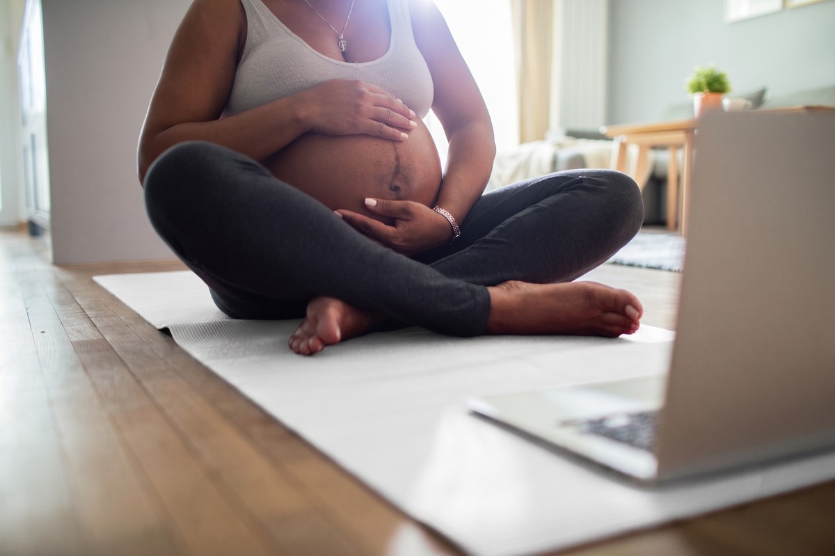 Pregnant woman sitting on yoga mat with laptop and baby bump.