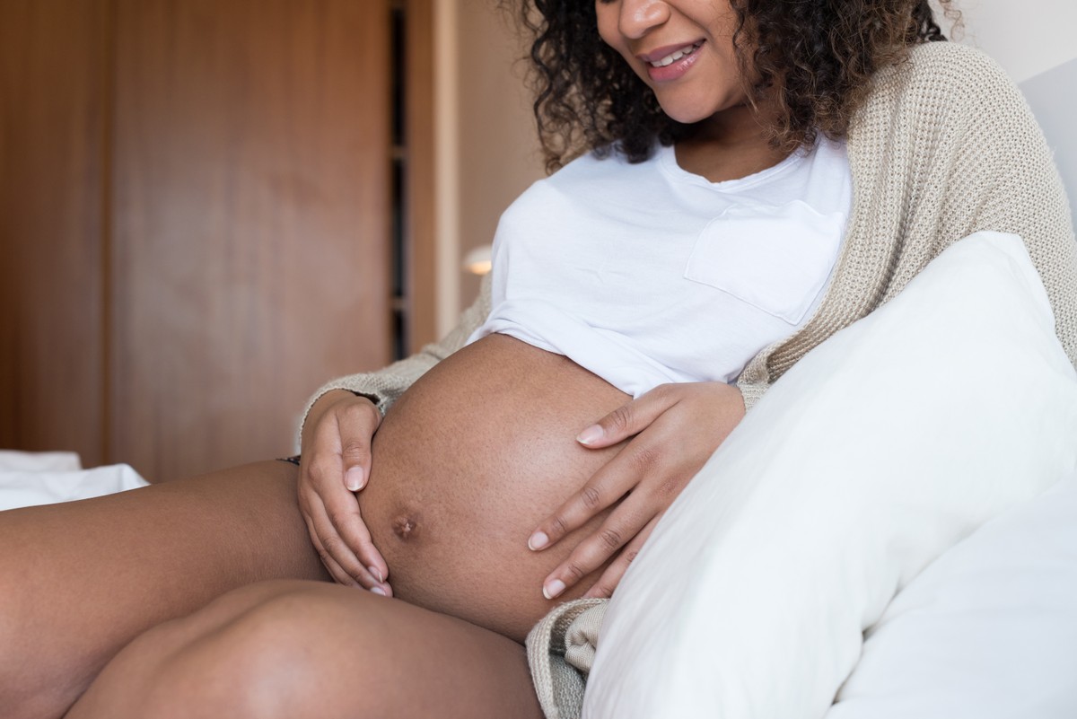 Pregnant woman holding a green bowl on counter, wearing white top, with various vegetables in the background.