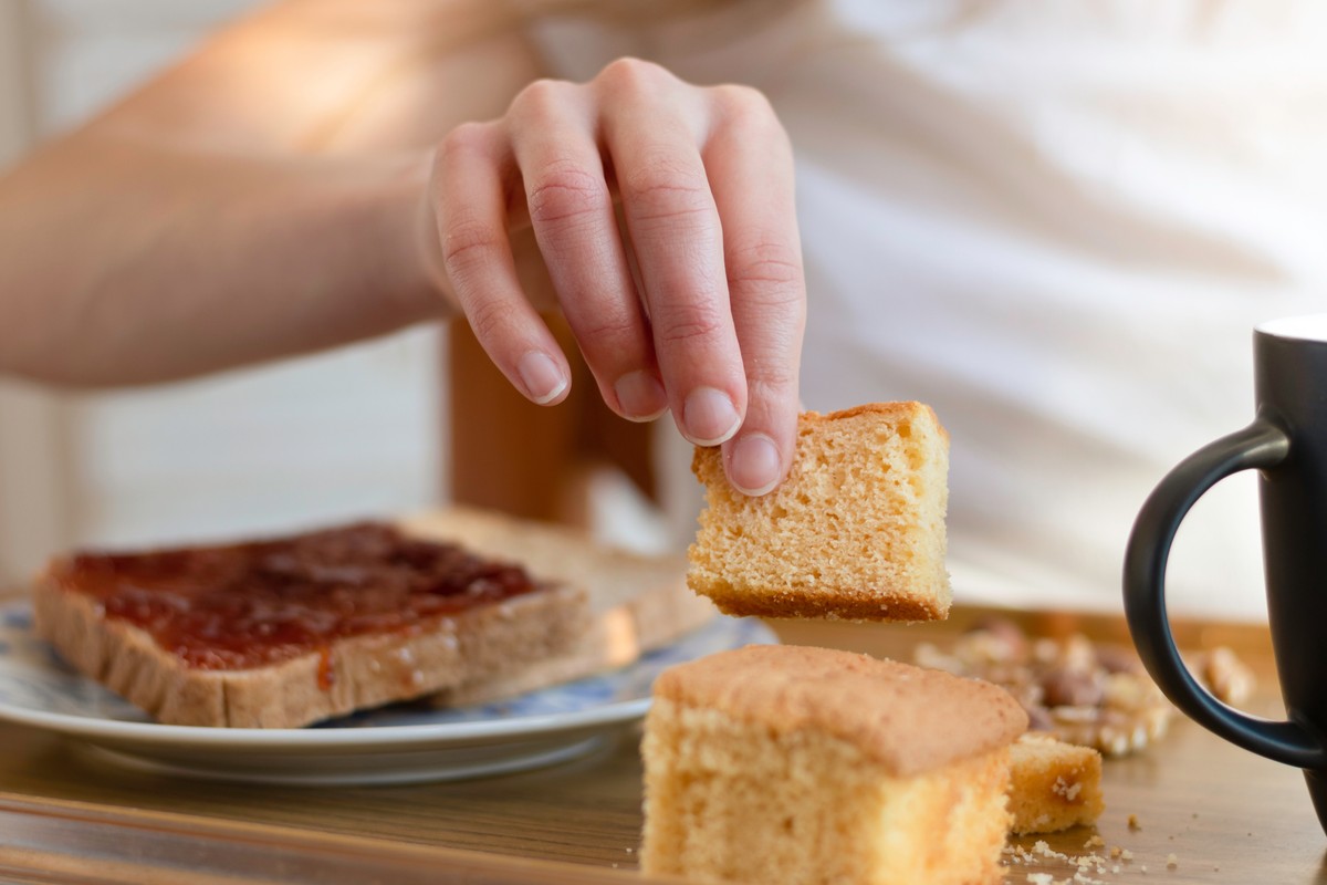 Hand holding a slice of cake in a room with wooden table and chairs.